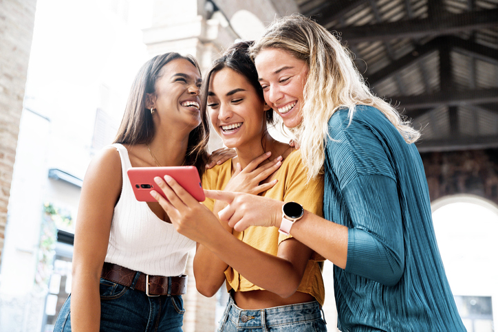 Three happy friends watching a smart phone mobile outdoors - Millennials women using cellphone on city street - Technology, social, friendship and youth concept