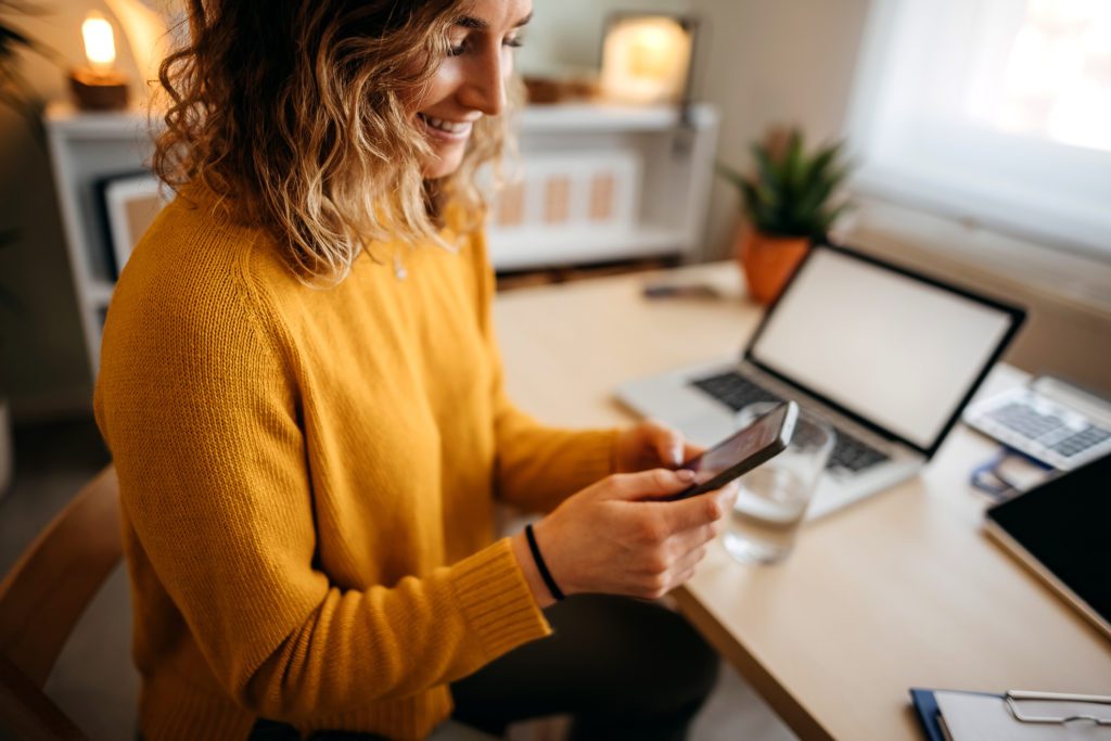 young woman sitting at the desk in a home office and using a smart phone to check email